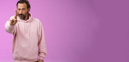 One rule listen. Serious-looking bossy focused determined adult bearded male in pink hoodie extend index finger first number scolding making statement forbid bad behaviour, purple background photo