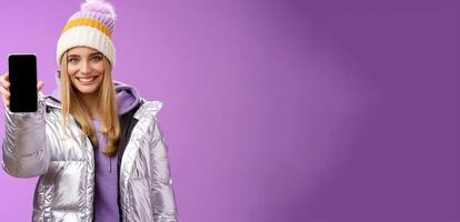 Friendly cheerful confident blond girl in silver stylish winter jacket hat extend arm showing smartphone display advertising awesome new device app smiling self-assured recommend use mobile phone photo