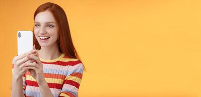 Redhead girl having fun recording hilarious friend actions hold smartphone look display amused shooting funny telephone standing orange background satisfied smiling delighted photo