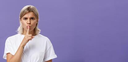Close-up shot of serious strict good-looking caucasian female with blond hair shushing at camera displeased and disappointed frowning holding index finger over folded lips in shush sign photo