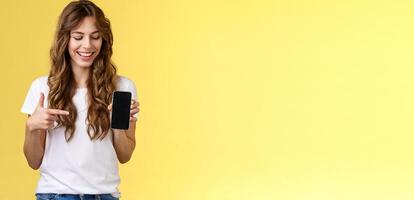 Intrigued charismatic happy young female blogger show what photo app use edit social media profile hold smartphone looking pointing index finger mobile phone blank screen smiling amused
