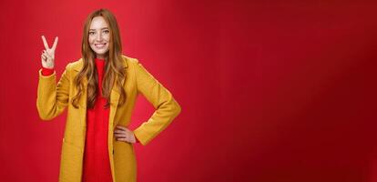 Portrait of happy playful and cute redhead woman with freckles in yellow coat holding hand on waist confident and self-assured showing peace or two sign delighted, beating weather conditions photo