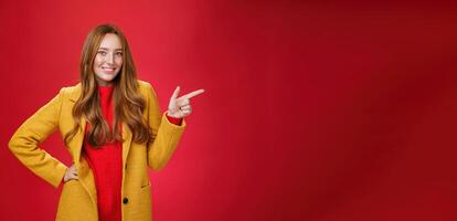 Stylish and self-assured attractive redhead woman with freckles in yellow outdoor coat pointing right with finger pistol and smiling joyfully as camera as showing cool promotion over red wall photo