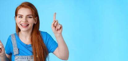 Close-up cheerful carefree redhead dancing girl pointing up, raising fingers, smiling amused, having fun, stand blue background entertained, enjoy music party, disco mood, carefree emotions concept photo