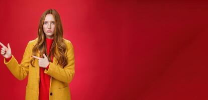 Displeased offended and gloomy girlfriend asking question, pouting and frowning from insult pointing at upper left corner disappointed and upset posing dissatisfied against red background photo