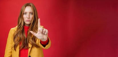 Serious-looking redhead woman in yellow coat extending hand to camera with stop and warning gesture prohibiting and forbidding make something having unemotional face as being confident over red wall photo