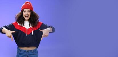 Hurry up and chek it out. Portrait of charismatic joyful and excited young cute curly-haired girl in beanie and stylish outfit pointing down, smiling broadly inviting join fun over blue background photo