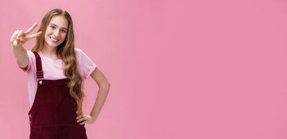 Charismatic young teenage girl in corduroy overalls posing joyfully and energetic against pink background showing peace sign at camera holding hand on waist in confident pose and smiling cheerfully photo