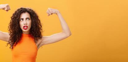 Woman feeling powerful and strong raising hands with clenched fists making intense face being working out in gym showing muscles and biceps looking at upper right corner posing over orange background photo