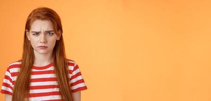 Close-up moody upset childish redhead girl, sulking unfair situation, frowning upset, complaining disappointed, pouting offended, stand uneasy perplexed orange background, whining displeased photo