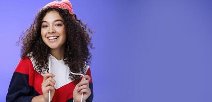Close-up shot of carefree attractive feminine girl with curly hair in beanie playing with sweatshirt as posing over blue background, smiling at camera having fun and positive attitude photo
