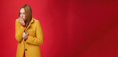 Girl getting cold, coughing and buttoning yelling light coat as freezing on chilly, windy weather looking displeased and ill at camera, feeling unwell catching flu over red background photo