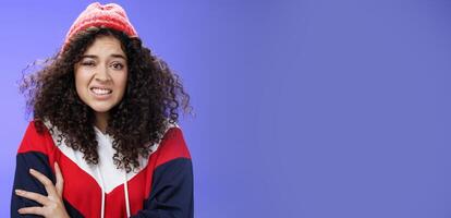 Woman with curly hair in winter beanie feeling uncomfortable and discomfort clenching teeth and frowning intense as hugging herself insecure and awkward, unwilling to say cruel rejection, feel awkward photo