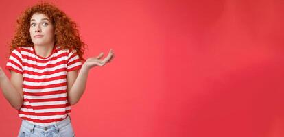 Not know ask someone else. Clueless unaware cute redhead curly-haired charming modern girl uncertain where spend summer holidays shrugging hands spread sideways smirking confused red background photo