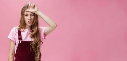 Waist-up shot of arrogant young girl with high ego grimacing snobbish showing letter L on forehead mocking over guy who lose calling person loser with scorn and disdain over pink background photo