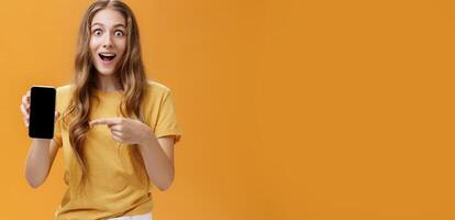 Girl surprised with awesome smartphone features. Portrait of delighted and amazed cute european slender woman with wavy natural hairstyle showing cellphone pointing at gadget screen at app photo