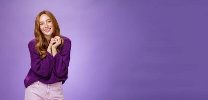 Charming and tender carefree redhead girlfriend with cute freckless holding hands together near shoulder as posing flirty and feminine smiling broadly having positive, happy attitude over purple wall photo