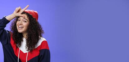 Girl triumphing and mocking friend as winning showing loser gesture on forehead and laughing having fun standing pleased and happy over blue background, smiling at camera joyfully photo