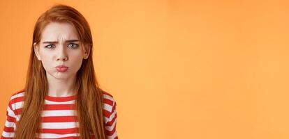 Childish offended cute redhead girl pouting, sulking upset, frowning, complain unfair situation, whining displeased, grimacing upset, act immature demand buy copy space orange background photo