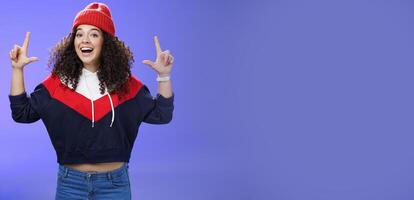 Waist-up shot of cheerful energized young woman calling friend inviting hang out as weather awesome pointing up with raised hands smiling broadly posing in red winter hat over blue background photo