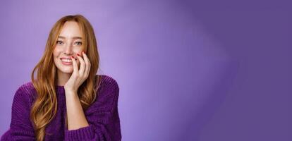 Close-up shot of feminine and glamour cute redhead girl with freckles in purple knitted sweater holding hand on cheek gently chuckling and smiling broadly at camera over violet background photo