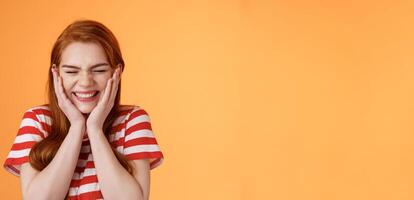 Close-up cheerful lucky excited smiling girl, cannot believe cheering hear excellent news, close eyes celebrating, touch cheeks blushing joy delight, rejoicing awesome situation, orange background photo