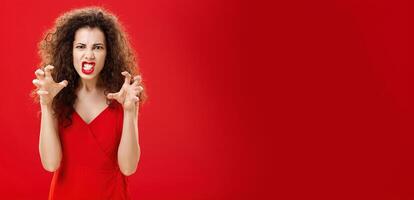 Woman like panter wanting scratch eyes of enemy. Portrait of pissed angry and pressured fed up woman clenching teeth and hands as if choking enemy with hate and anger in eyes being scary over red wall photo