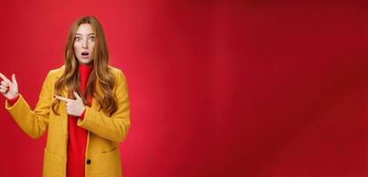 Stunned speechless and impressed attractive redhead woman with freckles in yellow coat drop jaw amazed as popping eyes at camera questioned, pointing left astonished over red background photo