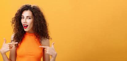 Proud and satisfied cool urban female with red lipstick and curly hairstyle pointing at herself with self-assured expression winking bragging about skills and achievements over orange background photo