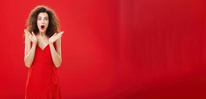 Woman learning surprising news being intrigued to hear all dirty ditails folding lips gasping making wow sound with raised palms in amazement gazing entertained and shocked at camera over red wall photo