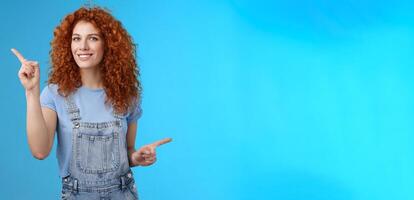 Lively joyful good-looking 25s redhead curly-haired sassy girl summer positive mood dancing overalls pointing sideways up right index fingers directing promos store links smiling delighted photo