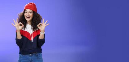 Got under control. Portrait of happy charming smiling curly-haired female in warm winter hat and sweatshirt smiling broadly and showing okay or excellent gesture as approving, liking cool movements photo