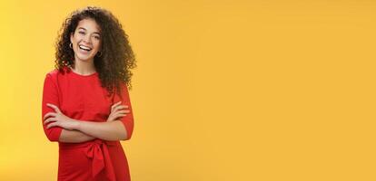 Self-assured happy enthusiastic curly-haired female reporter in cute red dress laughing carefree, having fun tilting head amused and holding hands crossed over body in confident pose over yellow wall photo