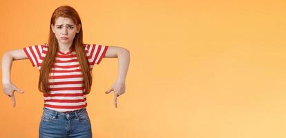 Cute timid hesitant ginger female taking tough decision feel pressured upset, frowning, pulling gloomy unhappy face, pointing down disappointed, uneasy taking decision, orange background photo