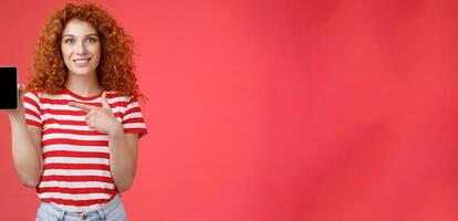 How about this app. Redhead curly friendly cute girl help friend find oufit online store hold smartphone pointing phone screen smiling broadly recommend social media photo filter, red background
