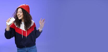 Attractive happy young woman with curly hair in hat singing enjoying perfect winter day singing along in smartphone, holding mobile phone like microphone, adore karaoke over blue background photo