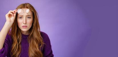 Waist-up shot of intense confused and unsure cute redhead female cannot look without glasses having bad sight taking off eyewear and squinting at camera uncertain, unable to read over purple wall photo