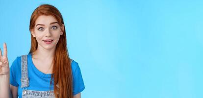 Surprised cute redhead caucasian girl show number three, third, counting fingers, raise eyebrows ambushed, smiling curiously, stand blue background playful tender expression photo