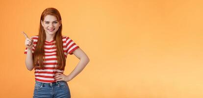 Cheerful sly redhead girlfriend have something interesting on mind, squinting cunning joyful smile, hold hand waist confident pose, pointing upper left corner give recommendation, orange background photo