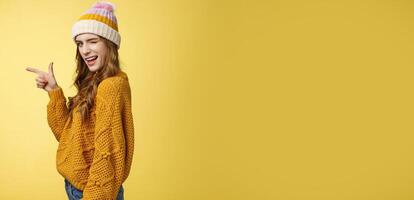 Profile shot sassy flirting young cute woman winking seduce you smiling cheeky turn camera pointing left index finger showing copy space suggest go drink coffee together, standing yellow background photo