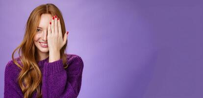 Fllirty and happy attractive ginger girl peeking with one eye as covering half face with palm, smiling dreamy from happiness and playful mood posing delighted and carefree over purple background photo