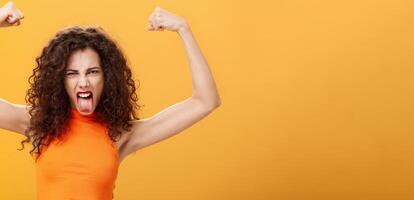Waist-up shot of cool and daring caucasian female in orange top with tattoo on arm frowning making funny face sticking out tongue raising hands showing muscles feeling power and strengths photo