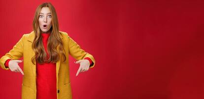 Impressed girl cannot believe what sees open mouth from surprise and excitement popping eyes questioned at camera, pointing down with index fingers asking question and being astonished over red wall photo