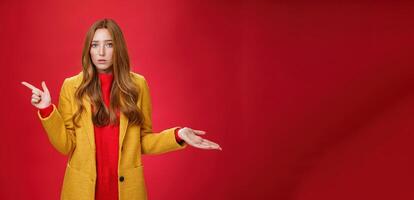 Frustrated and confused young cute female with ginger hair in yellow coat shrugging as pointing questioned left waiting explanation as asking question clueless and puzzled over red background photo