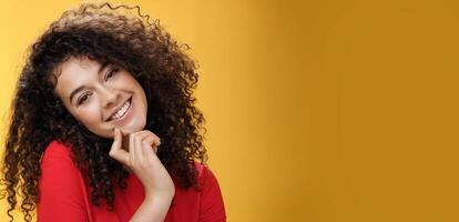 Close-up shot of charming flirty and silly curly-haired young woman making faces as trying get what wants, smiling tilting head on shoulder and touching lip standing cute over yellow background photo