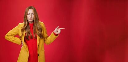 Explain yourself now. Portrait of upset gloomy and sad cute redhead girl waiting explanations asking question, pouting offended and pointing right with finger looking unhappy at camera over red wall photo