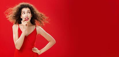 Waist-up shot of concerned and worried insecure cute female with curly hairstyle in red evening dress biting finger looking troubled or questioned at upper right corner with hand on waist photo