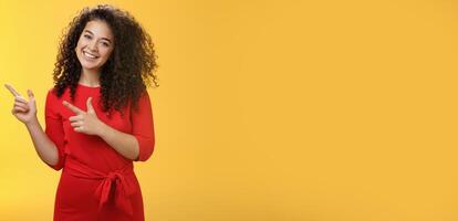 Friendly cute assertive female assistant in elegant red dress with curly hair tilting head flirty and smiling broadly, satisfied as pointing at upper left corner, recommending product over yellow wall photo