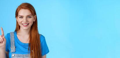 Close-up cute tender redhead european female make order, show number one first, smiling friendly carefree, extend finger, pointing up, counting stand blue background joyful photo