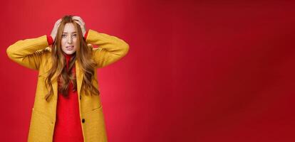 Worried and freaked out redhead girl holding hands on head grimacing troubled and concerned clenching teeth, frowning worrying and feeling nervous for project going fail over red background photo